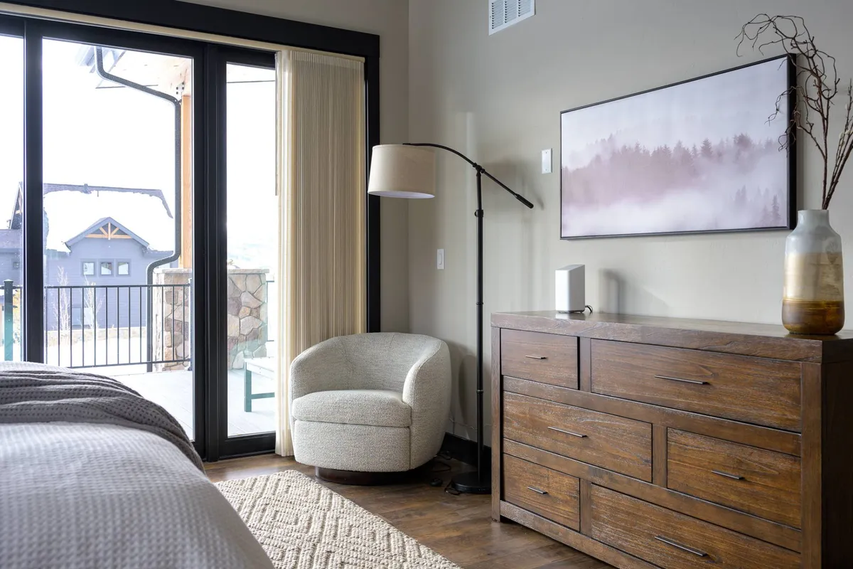 A serene interior space featuring large floor-to-ceiling windows that bring the outdoors in, a modern armchair positioned for enjoying the expansive mountain view, a wooden dresser, and framed artwork. The scene seamlessly connects the comfort of the indoors with the natural beauty just outside, suggesting a relaxing retreat after a day of adventures.