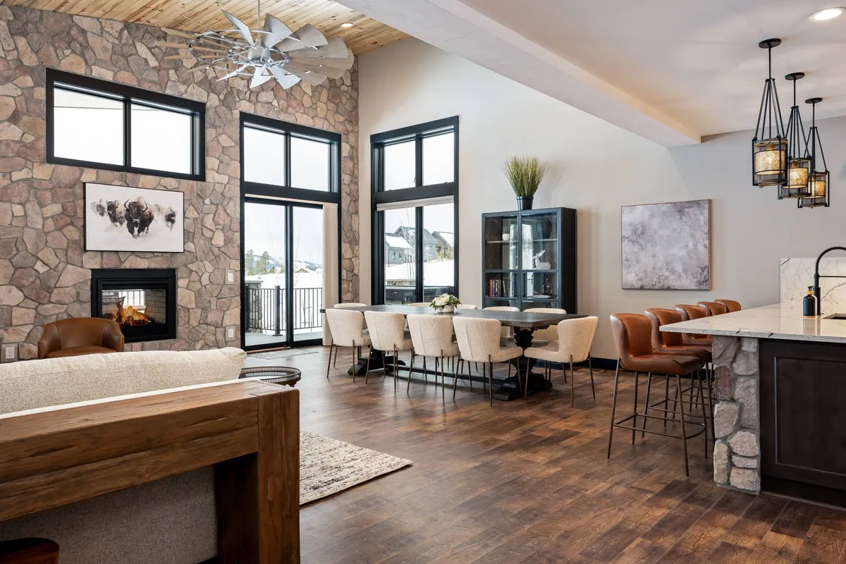 A spacious living and dining area highlighted by a beautiful stone fireplace, a large dining table, artful wall decor, and expansive windows that flood the space with natural light. The blend of stone, wood, and art creates a mountain-lodge ambiance perfect for family gatherings or festive celebrations.