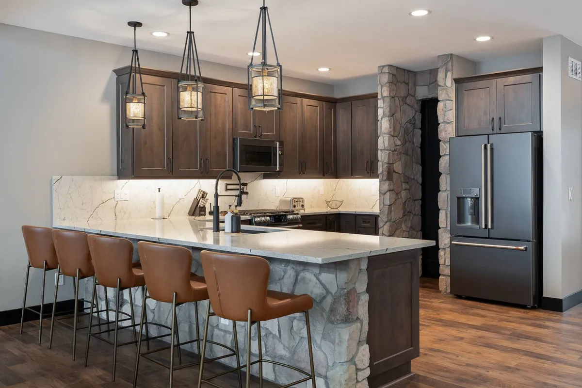 A well-appointed kitchen featuring a prominent island counter with bar stools, stylish hanging pendant lights, and a striking stone feature wall. Modern kitchen appliances are visible, suggesting a space ready for culinary creativity and entertaining. The scene feels both welcoming and functional, ideal for gathering and cooking together.