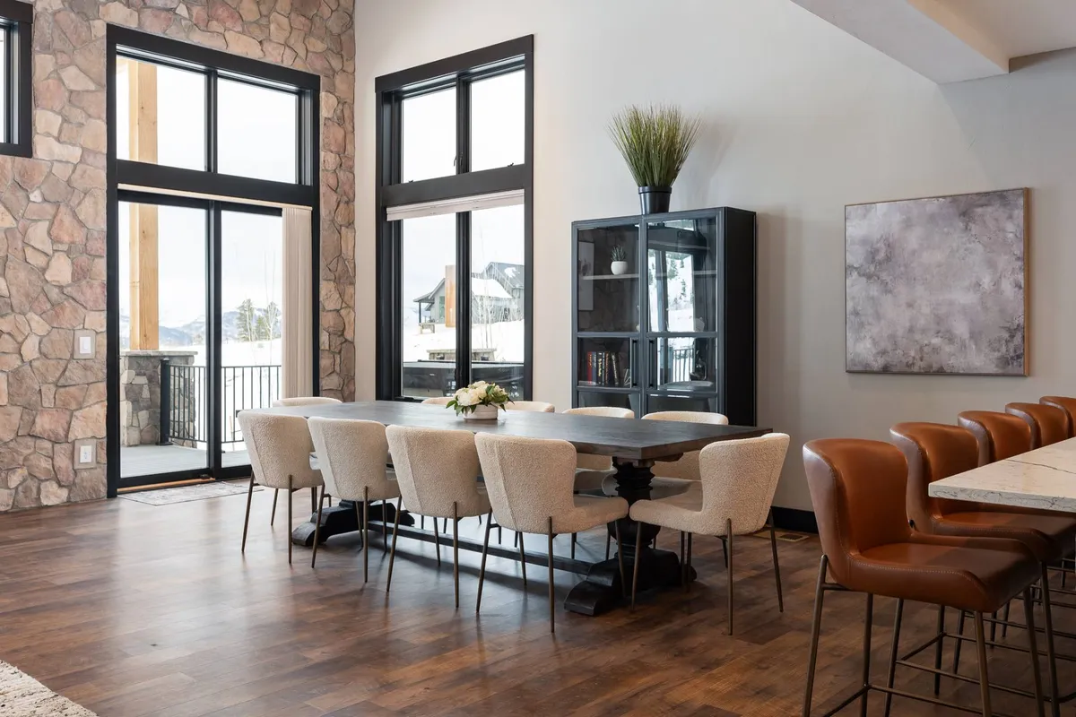 A warm and inviting dining area anchored by a large dining table, with expansive windows offering serene views, a stone wall accent, and adjacent bar stools. The atmosphere suggests an ideal space for family gatherings or holiday dinners.