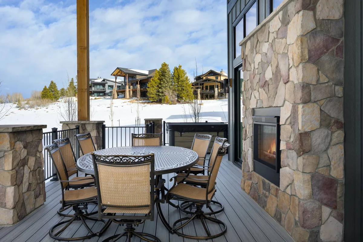 An exterior deck scene featuring a large stone fireplace, a dining table, and expansive views of snow-capped nature. People gather around the fireplace, enjoying the cozy warmth against the winter backdrop, suggesting après-ski relaxation and socializing outdoors.
