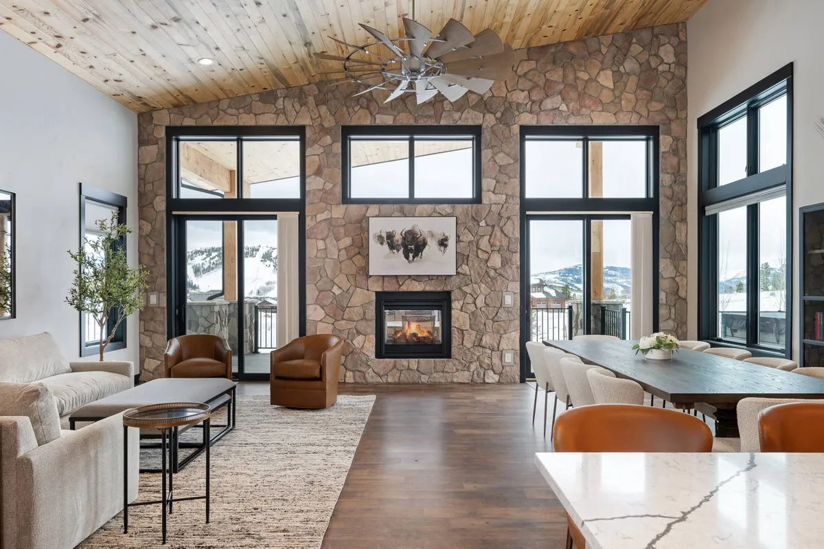 The image showcases the inviting interior of a luxury mountain home, centered around a grand stone fireplace. Expansive windows frame views of the surrounding landscape, filling the space with natural light. A spacious dining table and plush, comfortable seating create the perfect setting for family gatherings, cozy evenings, and memorable meals.